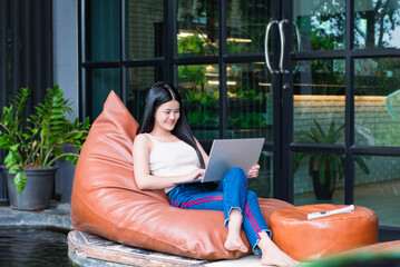 Young adult asian businesswoman sitting on orange pillow, smiling and looking down to her laptop in front of glassdoor of the cafe during her long vacation, digital nomad of woman concept.