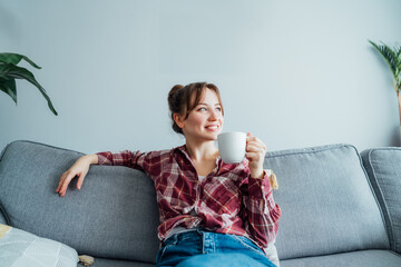Young smiling woman sitting on sofa and looking side up while drinking coffee or tea. Young brunette woman relaxing after housekeeping, home cleaning. Portrait of relaxed female resting at home