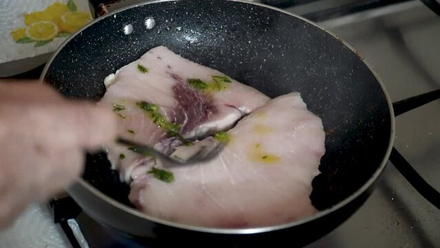 A person seasons with oil and parsley some slices of swordfish frying in the pan.