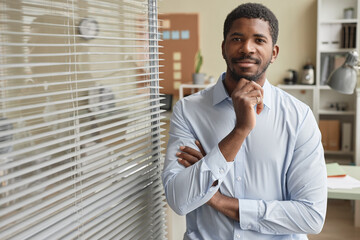 Waist up portrait of smiling black businessman by glass wall in office looking at camera with hand on chin, copy space