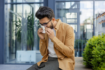 Young man sneezing and having a runny nose allergy sitting on a bench in the daytime outside an...