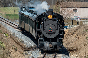 Naklejka premium A Front and Slightly Above View of an Approaching Restored Narrow Gauge Passenger Steam Train Blowing Smoke and Steam on a Sunny Winter Day