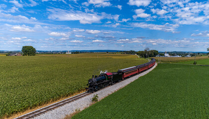 A Drone Frontal View of a Restored Steam Passenger Train, Traveling Thru Green Corn Fields, Blowing Smoke on a Fall Day