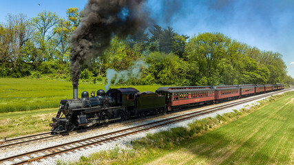 Obraz premium An Aerial View from the Side of An Antique Steam Locomotive and Passenger Coach Stopped and Blowing Smoke and Steam, While Waiting for Passengers to Board on a Sunny Spring Day
