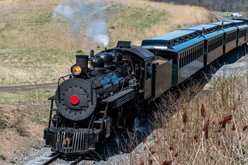 Obraz premium A Front and Slightly Above View of an Approaching Restored Narrow Gauge Passenger Steam Train Blowing Smoke and Steam on a Sunny Winter Day