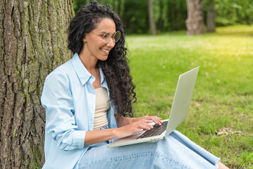 Portrait of a young Middle Eastern woman with long curly hair sitting on the grass near a tree and using a netbook while doing her homework on a summer day in the park. Busy girl doing work with enjoy