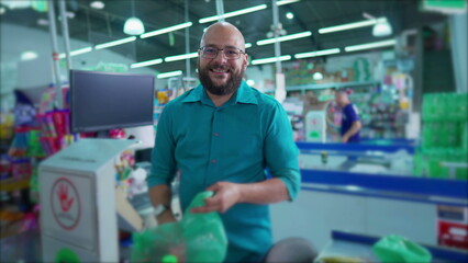 Naklejka na ściany i meble One happy Brazilian employee of supermarket at checkout scanning products and putting into plastic bags while smiling at camera. Grocery Market scene in South America