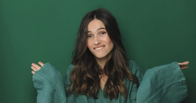 Puzzled confused brunette young woman wearing green dress posing isolated on green color background in studio. Curly hair brunette woman looking camera spreading hands say oops.