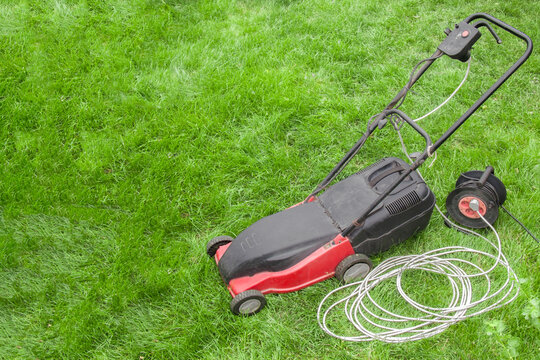 red electric lawn mower on a green lawn with wires and an electric extension cord, portable socket