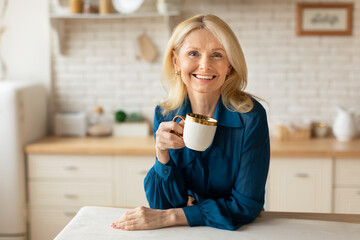 Cheerful mature woman enjoying herbal tea at home, standing at cozy kitchen, holding coffee mug and smiling at camera