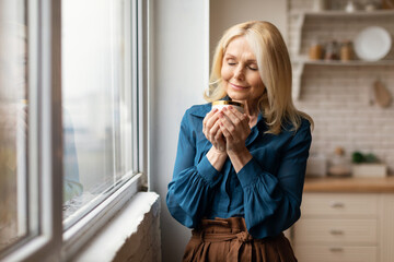 Cheerful mature woman enjoying morning coffee, holding mug in hands and smelling hot drink with closed eyes in kitchen