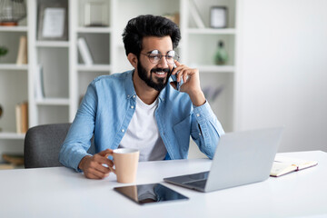 Remote Business. Smiling Male Indian Freelancer Talking On Cellphone And Using Laptop