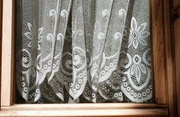Fenster mit braunem alten Holzrahmen und grau-weißen Gardinen mit Blumenmuster hinter Glasscheibe bei Sonne am Nachmittag im Sommer