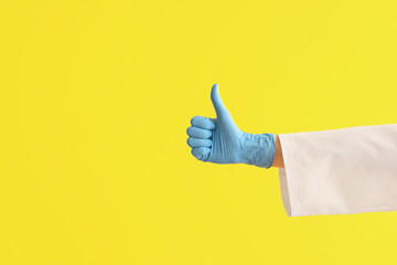 Female doctor's hand in medical glove showing thumb-up on yellow background