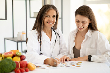 Smiling mature caucasian doctor nutritionist in white coat consultation young lady at table with organic vegetables