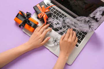 Female hands with modern laptop, gift boxes and web for Halloween on purple background