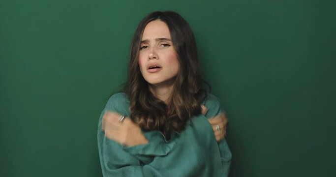 Young woman freezes in dress warms up by hugging herself, seasonal changes low temperature heating problem, looks at camera, chills feeling of coldness concept, isolated on green background.