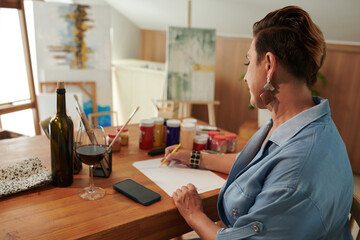 Mature woman drinking wine when drawing pictures at desk in her studio