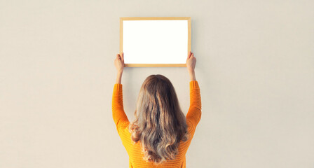 Young woman decorating interior, hanging blank a photo frame mockup on white wall in a new house - obrazy, fototapety, plakaty