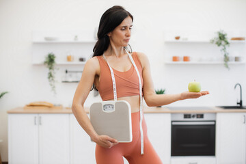 Fototapeta na wymiar Slender caucasian woman with measuring tape and scales holding apple on palm while standing in kitchen. Effective health coach in sportswear assisting in weight management via healthy nutrition.