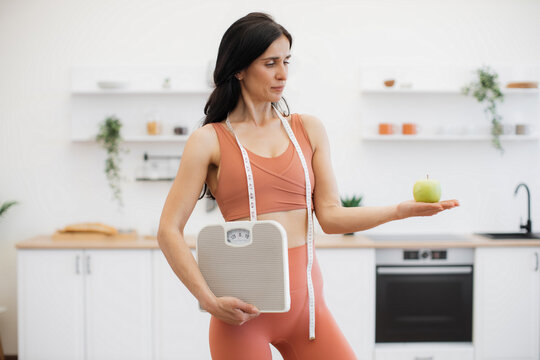 Slender caucasian woman with measuring tape and scales holding apple on palm while standing in kitchen. Effective health coach in sportswear assisting in weight management via healthy nutrition.