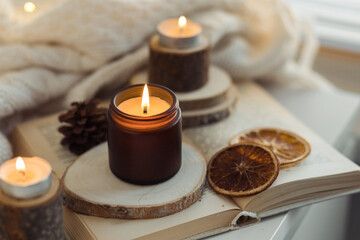 Cozy autumn or winter composition with aromatic candle, wool sweater, fairy lights, book. Aromatherapy, home atmosphere of cosiness and relax. Wooden background close up