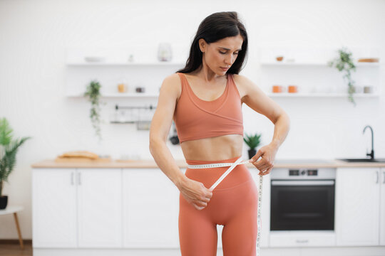 Attractive Woman In Good Health Measuring Waist With Tape After Working Out At Home In Yoga Activewear. Athletic Person Steering In Direction Of Improving Well-being Via Food Habits And Fitness.