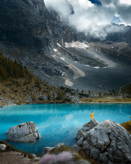 Beautiful mountain landscape with a light blue or blue lake and a man in a yellow raincoat. Beautiful mountains with fog or clouds around, Dolomites, Italy, Lago di Sorapis. Famous lake.
