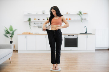 Amazed brunette female staring at loose pants on slim figure while standing on scales in middle of modern kitchen. Happy woman feeling pleasant shock of attained success in nutritional therapy.