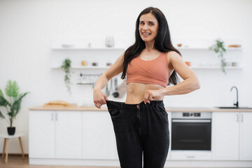 Surprised lady in terracotta top looking at black oversized jeans on thin waist while standing in kitchen of apartment. Sporty slim woman enjoying amazing achievements of well-balanced diet at home.