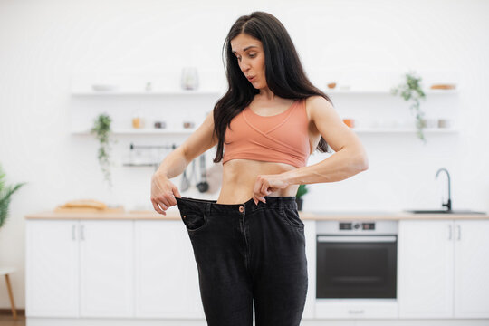 Surprised lady in terracotta top looking at black oversized jeans on thin waist while standing in kitchen of apartment. Sporty slim woman enjoying amazing achievements of well-balanced diet at home.