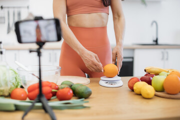 Close up view of slender woman weighing orange on smart digital scale while gadget on tripod shooting the process. Lady in good shape giving advice on improving diet with low-calorie products.