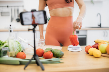 Obraz na płótnie Canvas Focus on female in activewear checking nutrition value of sweet pepper via scales in home kitchen. Sports-loving food influencer filming dietary advice about benefits of organic products on phone.