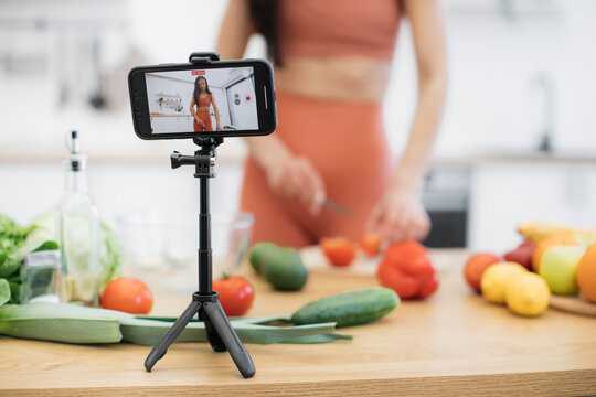 Cropped view of brunette female slicing tomato on cutting board and getting record of process on mobile. Athletic adult in activewear blogging about meal modificatons via vegetable rich in nutrients.