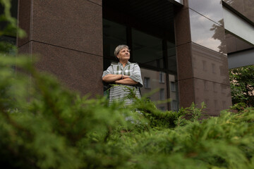 Real Happy Smiling Senior Woman In Yard Of Modern Building. Green Bushes Around. Copy Space For...