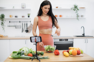 Beautiful young woman in sportswear adding oil to vegan salad on webcam of smartphone fixed by tripod in kitchen. Fit and healthy adult recommending fresh ingredients for health blog subscribers.