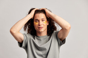 Young handsome long-haired guy applying under eye hydrogel patches on his face. Caucasian brown-haired millennial man practices skin care routine to keep healthy and youthful looking. Studio portrait.