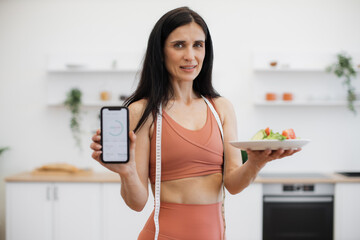 Portrait of motivated fitness woman posing on camera with smartphone and salad bowl while staying...