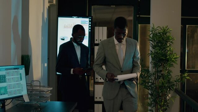 Supervisor debriefing assistant in front of digital screen, showing him key statistical data before getting back to work. Staff members in office during nightshift crosschecking business insights