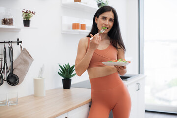 Pensive fitness person dressed in gym clothes standing in kitchen and looking at plate with greens. Long-haired brunette promoting healthy lifestyle with organic balanced food and regular training.