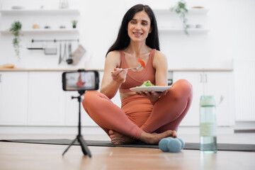 Female fitness blogger creating video content for social media about healthy dieting using tripod with smartphone. Fit lady sitting on floor of modern kitchen and eating fresh vegetable salad.