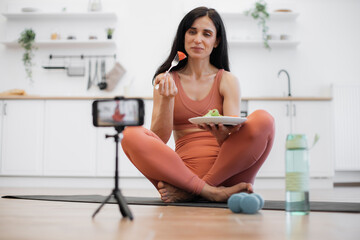 Beautiful dark-haired fitness coach recording video on cellphone telling about healthy and junk food. Sporty female taking break after workout for having snack. Blogging activity concept.