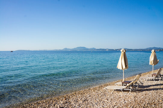 View of empty beach - white deck chairs and umbrellas near sea water, pebbles, mountains on the horizon and blue sky. Landscape of abandoned summer resort. Barbati, Corfu, Greece