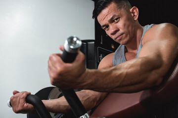 Fototapeta na wymiar A muscular and composed asian man looking at the camera before doing machine preacher curls at the gym. Arm and bicep workout training.