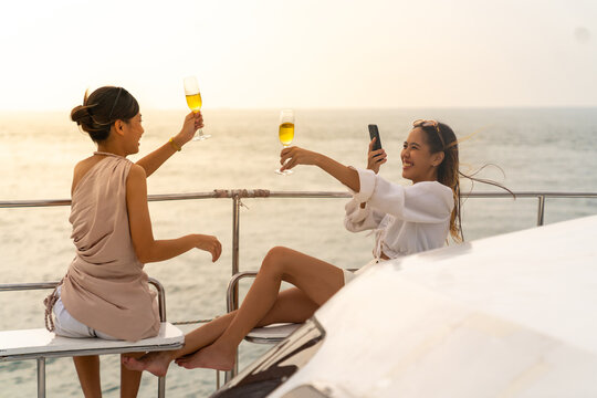 Young Asian woman friends using digital smartphone taking picture together during travel ocean on luxury private catamaran boat yacht sailing in the sea on summer holiday vacation at tropical island.