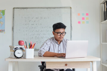 Young focused Asian male teacher or lecturer wearing glasses working on laptop. Remote studying using computer looking at screen, watching seminar webinar at home office.