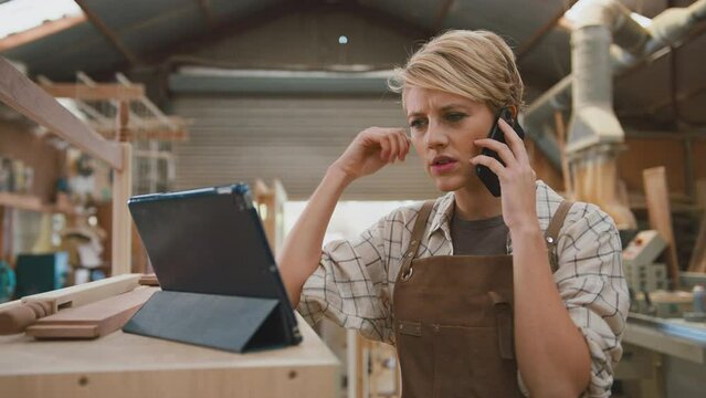 Female apprentice carpenter in furniture workshop using digital tablet and talking on mobile phone - shot in slow motion