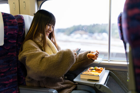 Asian woman using mobile phone taking picture of traditional Japanese lunch box bento during travel on train. Attractive girl travel Japan on railroad transportation on winter holiday vacation.