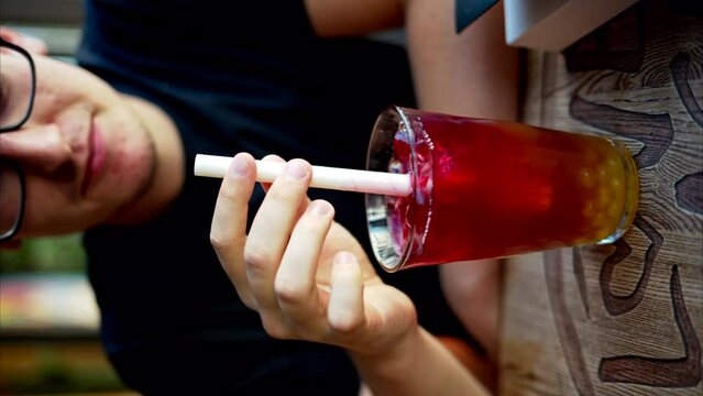 Man drinking red bubble tea with boba in a glass with ice and big white straw close up slow motion, vertical screen