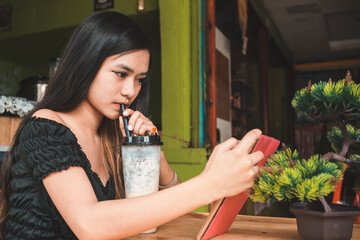 A young Filipina woman reads an ebook on her tablet while sipping on Milk tea at a small cafe.